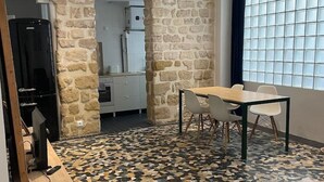 A room with a stone wall, a tiled floor, a dining table with chairs, a television, and a window with blinds. - Luma Oberkampf Apartment / Jean Pierre Timbaud (Paris)