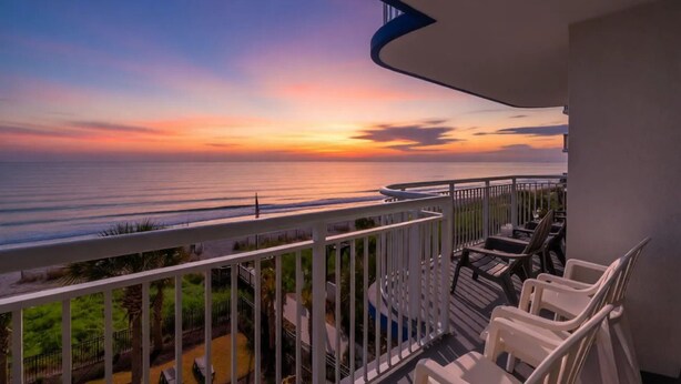 Un balcón con vistas al océano al atardecer, amueblado con mobiliario exterior.