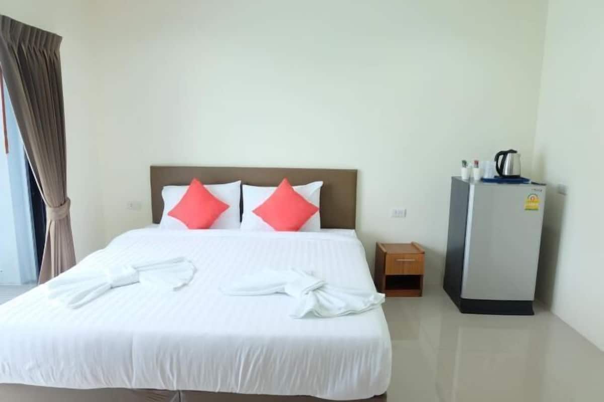 Una habitación de hotel con cama, almohadas, mesita de noche, refrigerador y ventana con cortinas.