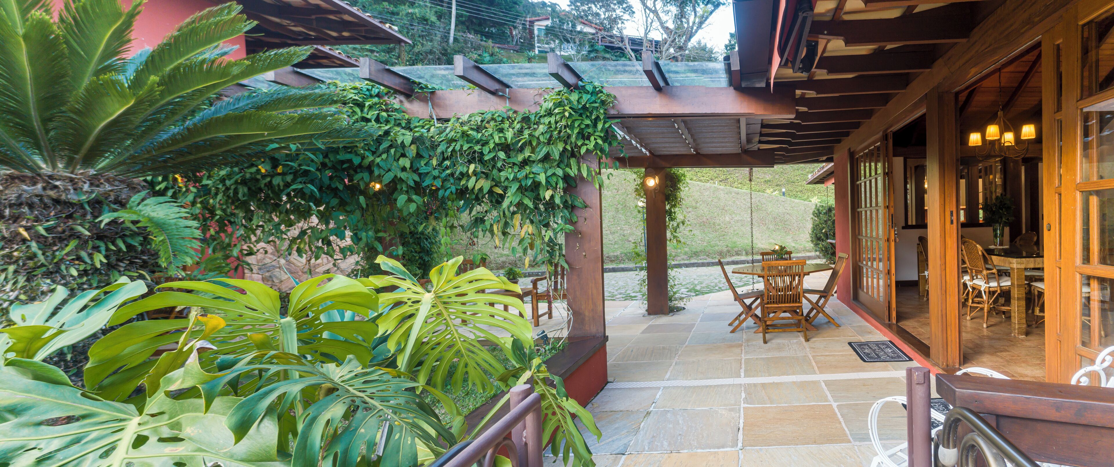 Une terrasse couverte, agréée d’une table et de chaises, entourée d’une végétation luxuriante et d’une pergola en bois.