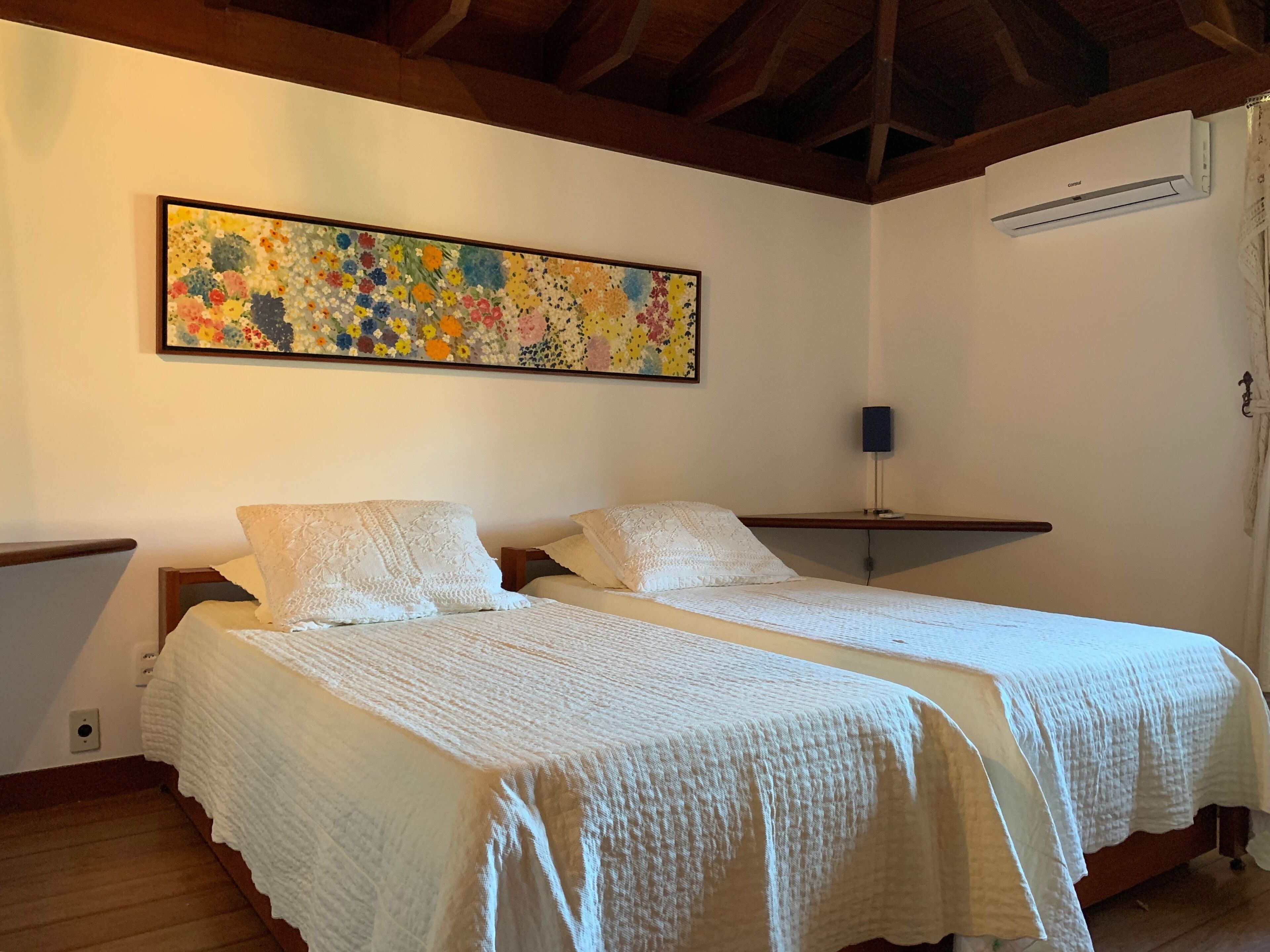 A room with two beds, a ceiling fan, a wall-mounted air conditioner, and a colorful abstract painting.