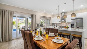 A modern kitchen with a dining area, featuring a wooden table set for four, a large island with bar stools, and a view of the outdoors. - Amazing 4BR 3BA Villa Palm Beach w NEW Pool (Noord)