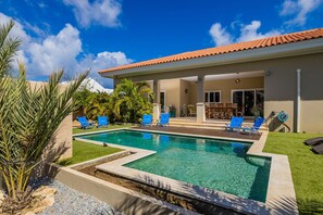 Une piscine rectangulaire bordée d’une terrasse carrelée, entourée d’un petit muret en pierre. Quatre chaises longues bleues sont alignées le long du bord de la piscine.