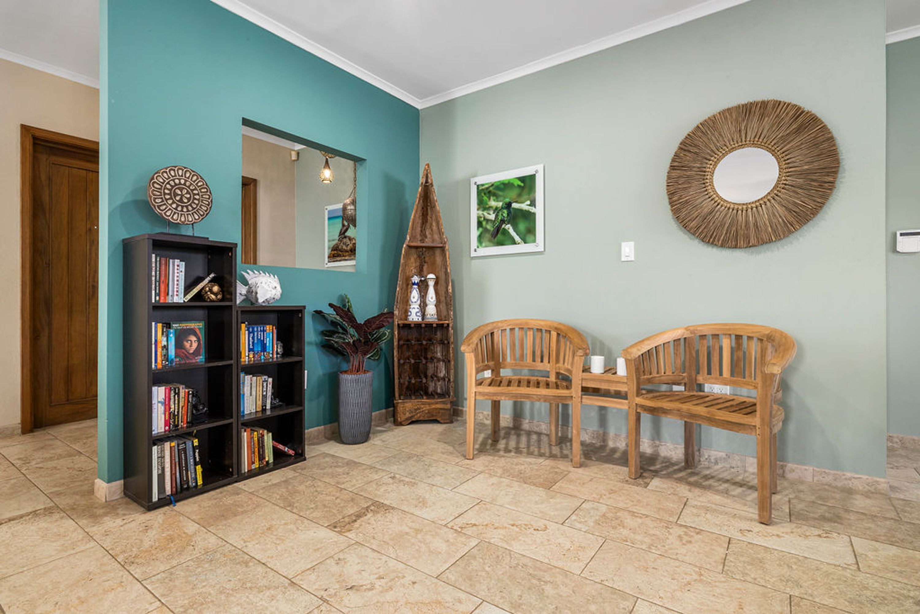 A room with teal walls, a wooden bookshelf, two wooden chairs, a mirror, and a decorative woven wall hanging.