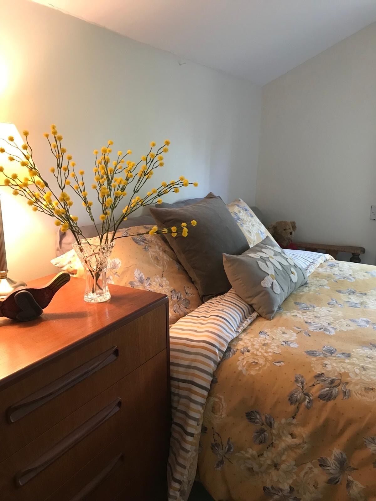A bedroom with a wooden dresser, a bed with floral bedding, and a vase with yellow flowers on the dresser.