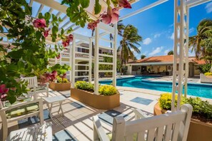 Un área junto a la piscina con sillas blancas y macetas, una piscina azul cristalina y un edificio con balcones al fondo.