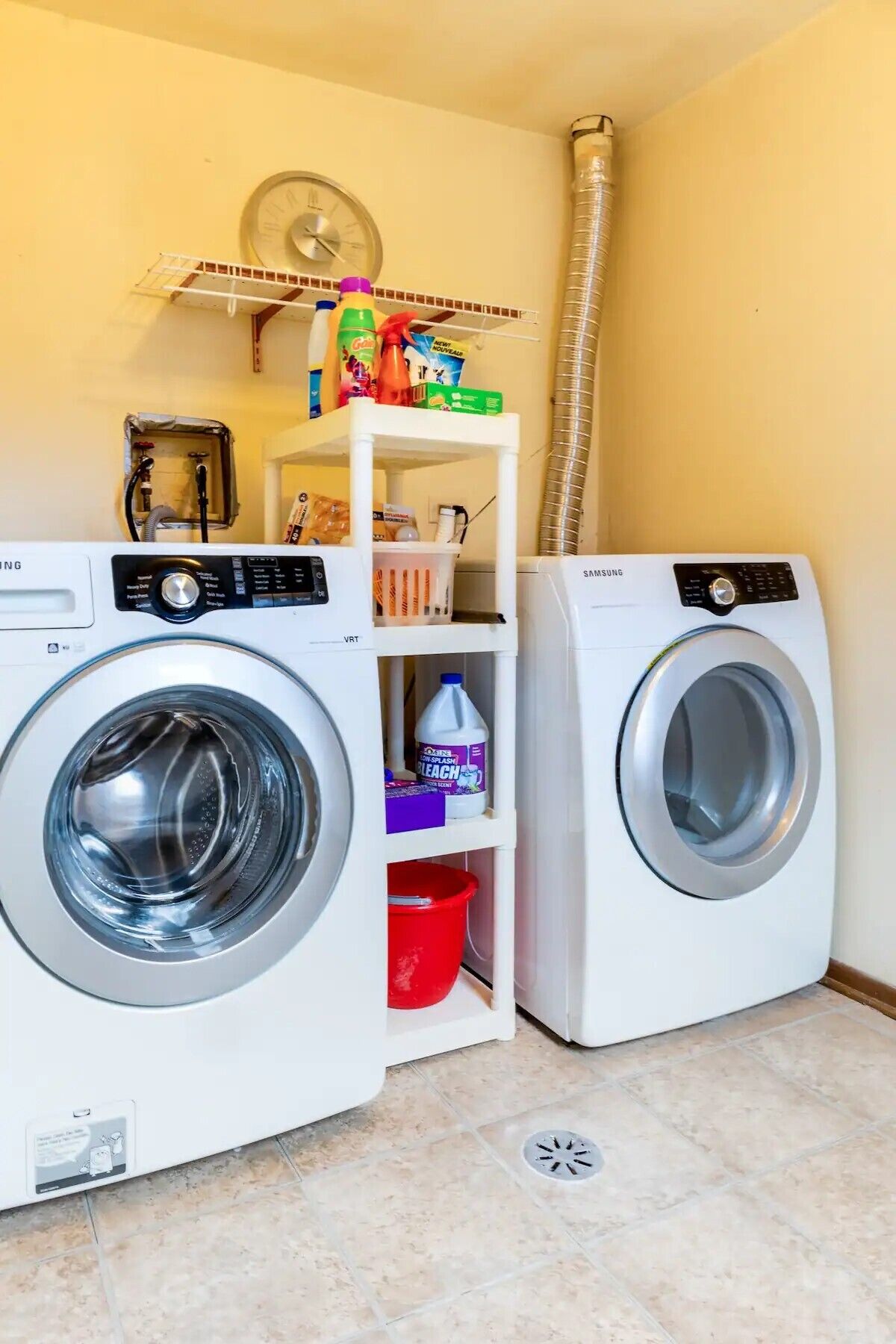 A laundry area with two front-loading washing machines, a shelf with cleaning supplies, and a vent pipe.