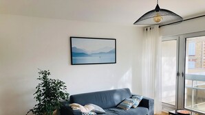Un salon moderne avec un canapé gris, une petite table d’appoint, une plante en pot et une grande fenêtre donnant sur des immeubles.