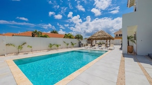 A rectangular swimming pool with a clear blue water, surrounded by a white tiled deck. There are lounge chairs and a thatched-roof structure in the background. - Buddha House 5 5 Palapa Suite Pool in Palm Beach (Noord)