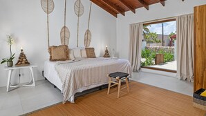 A bedroom with a bed, a small stool, a nightstand with a plant, and a window with curtains. - Buddha House 5 5 Palapa Suite Pool in Palm Beach (Noord)