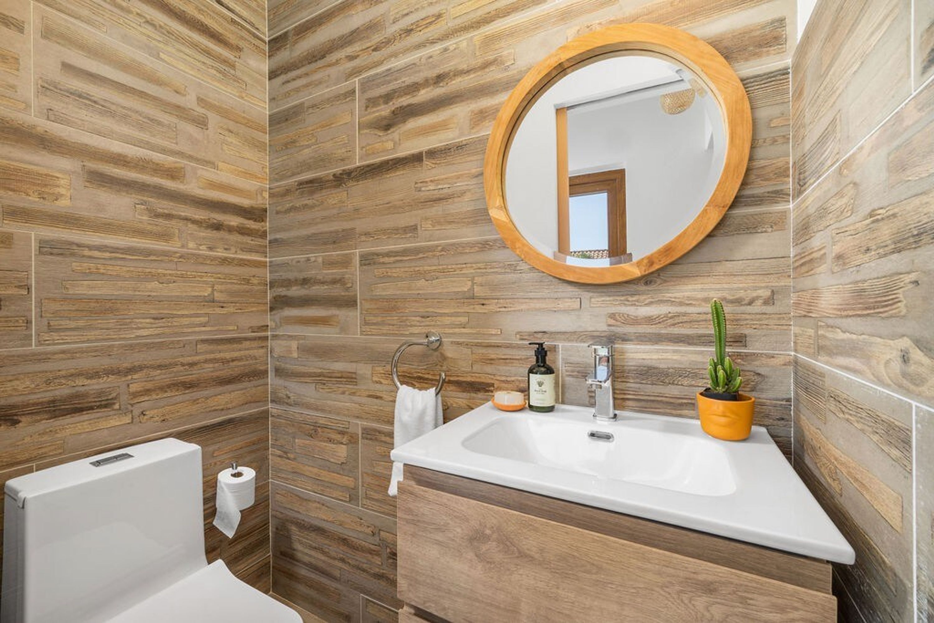 A bathroom with a wooden wall design, a round mirror, a sink, a toilet, and a small potted plant.