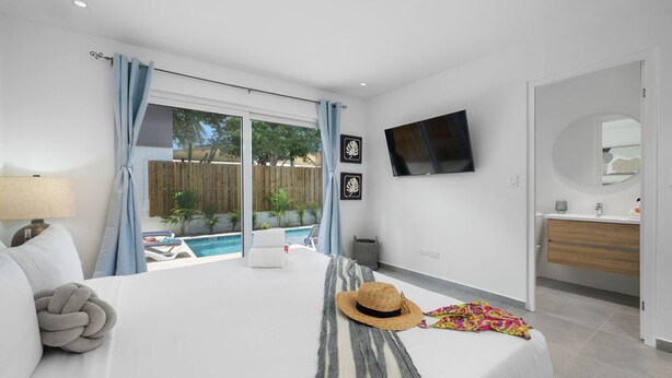 A bedroom with a bed, a TV mounted on the wall, a window with curtains, and a view of a swimming pool outside. - Brand NEW Private Pool King Beds Palmbeach (Noord)