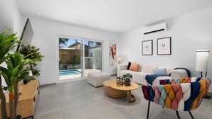 A modern living room with a sofa, coffee table, and colorful armchair. There is a TV on a stand, a plant, and a sliding glass door leading to a pool. - Brand NEW Private Pool King Beds Palmbeach (Noord)