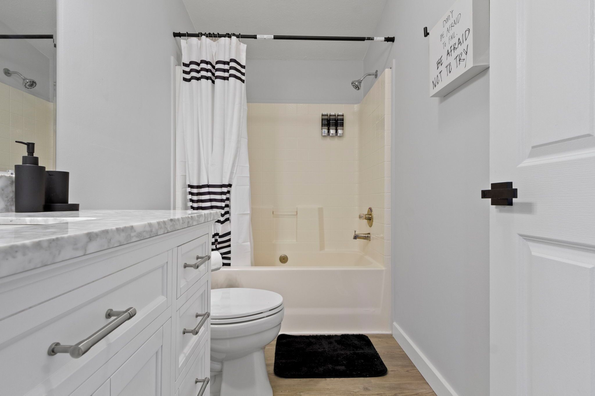 A modern bathroom with a white toilet, a bathtub, and a shower. There is a black and white striped shower curtain, a black rug, and a sign on the wall.