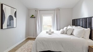 Ein Schlafzimmer mit einem großen Bett, weißer Bettwäsche, einem Kopfteil, einem Fenster mit Vorhängen und einem an der Wand hängenden Kunstwerk.