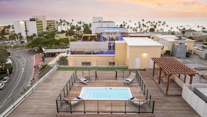A rooftop pool area with lounge chairs, a pool, and a view of the ocean and palm trees.