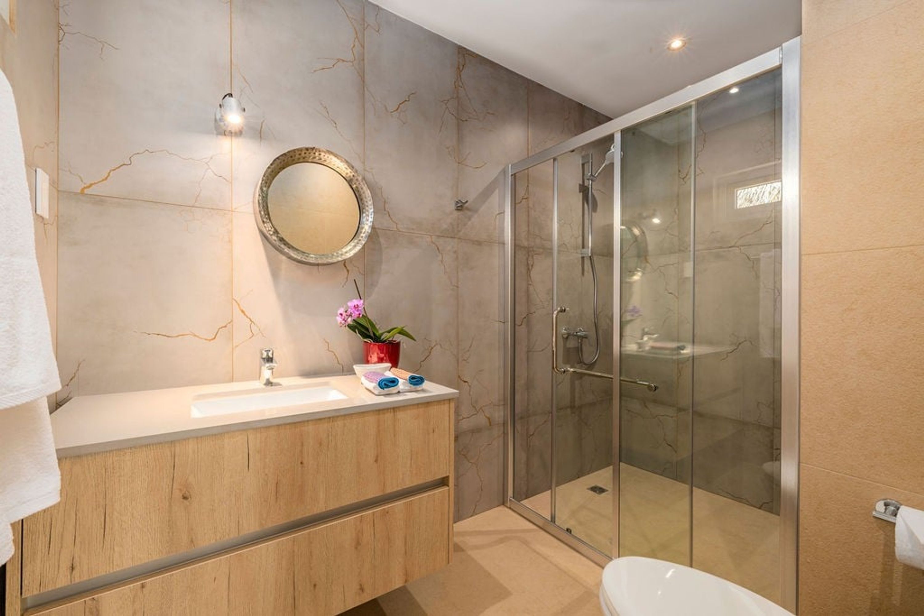 Un baño moderno con pared de mármol, tocador de madera, cabina de ducha de vidrio y bañera blanca.