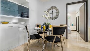 Une cuisine moderne avec des meubles blancs, un coin repas avec deux chaises noires, une horloge murale ronde et un couloir menant à une autre pièce.