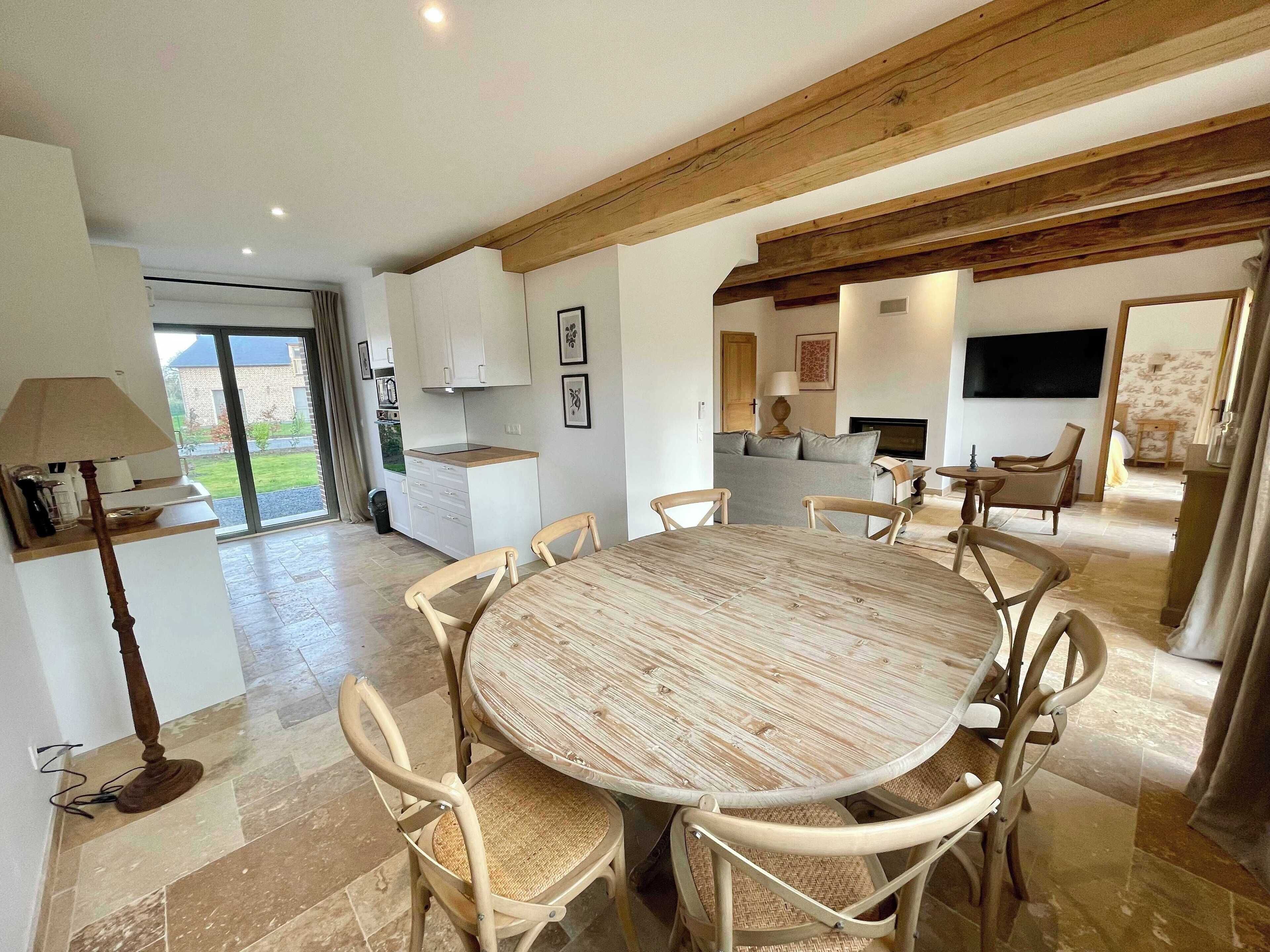 Un vaste espace de salle à manger avec une table ronde en bois et six chaises, une cuisine dotée de meubles blancs et une grande fenêtre donnant sur un jardin.