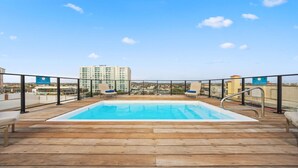 A rooftop pool with wooden decking and metal railings, overlooking a cityscape with high-rise buildings. - Lux Palm Beach Escape Pool Gym Prime Location (Noord)