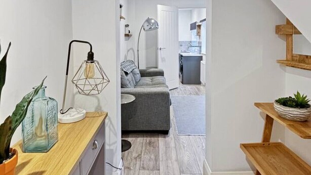 A narrow hallway leading to a living area with a grey sofa, a wooden bench, and a potted plant.