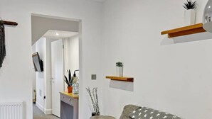 A modern living room with a grey sofa, wooden flooring, white walls, and a kitchen in the background.