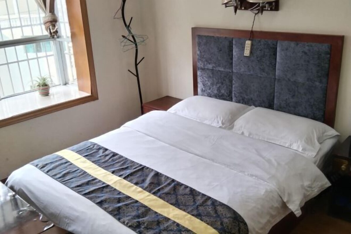 Una cama con cabecera, una mesita de noche, una ventana con planta y una lámpara fijada a la pared.