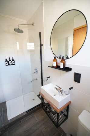 A modern bathroom with a glass-enclosed shower, a white sink with a wooden countertop, and a round mirror above the sink. - Güer Aike Tiny Houses (El Chalten)