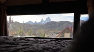 A tented room with a bed and a view of a mountainous landscape and a small building.