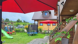 A wooden house with a sloped roof, a trampoline, a swing set, and a red umbrella.
