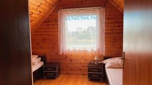 A cozy bedroom with wooden walls, a window with lace curtains, two bedside tables, and a lamp.
