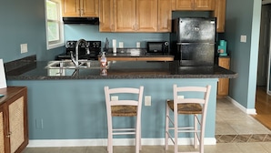 A kitchen with teal walls, wooden cabinets, a black refrigerator, a microwave, and a sink. There are two pink chairs at the island.