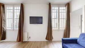 Een kamer met een wandgemonteerde flatscreen televisie, twee ramen met bruine gordijnen, een radiator en een blauwe bank.