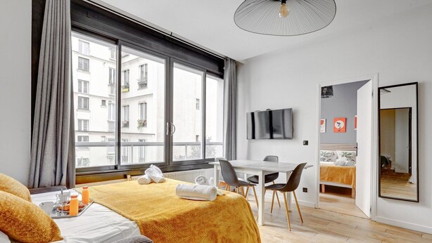 Et hotelværelse med seng, spisebord, fjernsyn og et stort vindue med udsigt til bygninger.