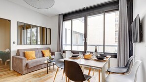 En moderne stue med sofa, spisebord og stole. Der er et stort vindue med gardiner, et vægmonteret fladskærms-tv og trægulv.
