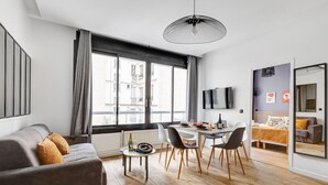 En moderne stue med sofa, spisebord og stole, store vinduer med gardiner, et tv ophængt på væggen, og en seng i det tilstødende rum.