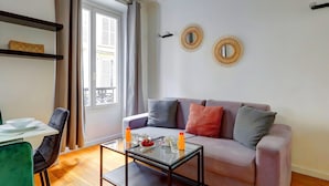 Ein Wohnzimmer mit einer Couch, einem Glastisch, einem Esstisch mit Stühlen und einem Fenster mit Vorhängen.