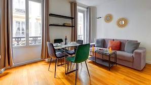 Ein modernes Wohnzimmer mit Essbereich, einer Couch und einem Couchtisch. Zwei Fenster mit Vorhängen, ein Bücherregal und dekorative Spiegel sind an der Wand angebracht.