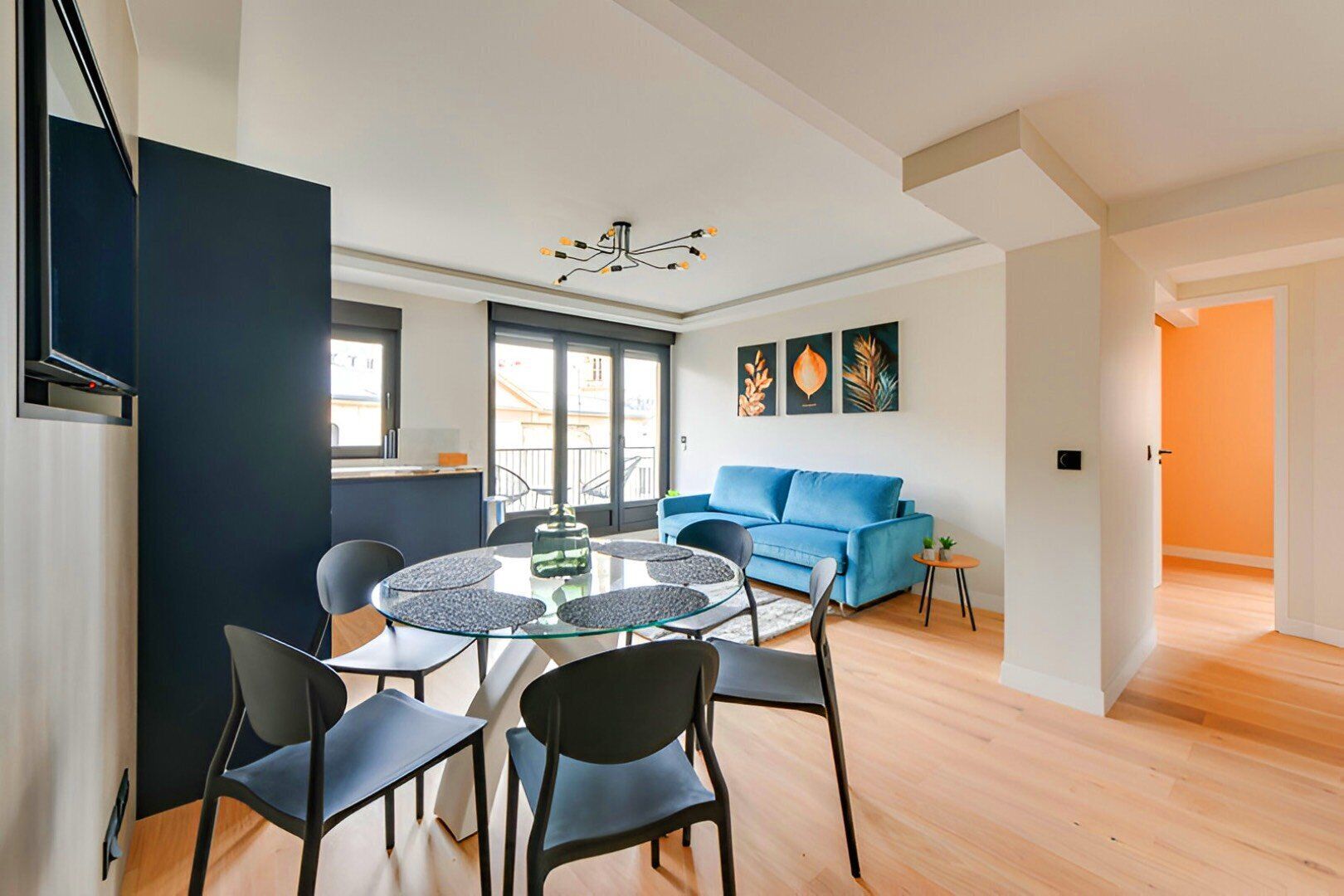 A modern living room with a round glass table, black chairs, a blue sofa, and a ceiling light fixture.