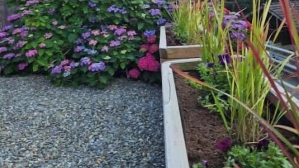 Un rustico chalet in legno con un vialetto sterrato che conduce a un'aiuola con fiori viola.