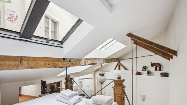 Una habitación espaciosa con techos altos, tragaluces, vigas de madera a la vista y una cama con toallas blancas ordenadas.