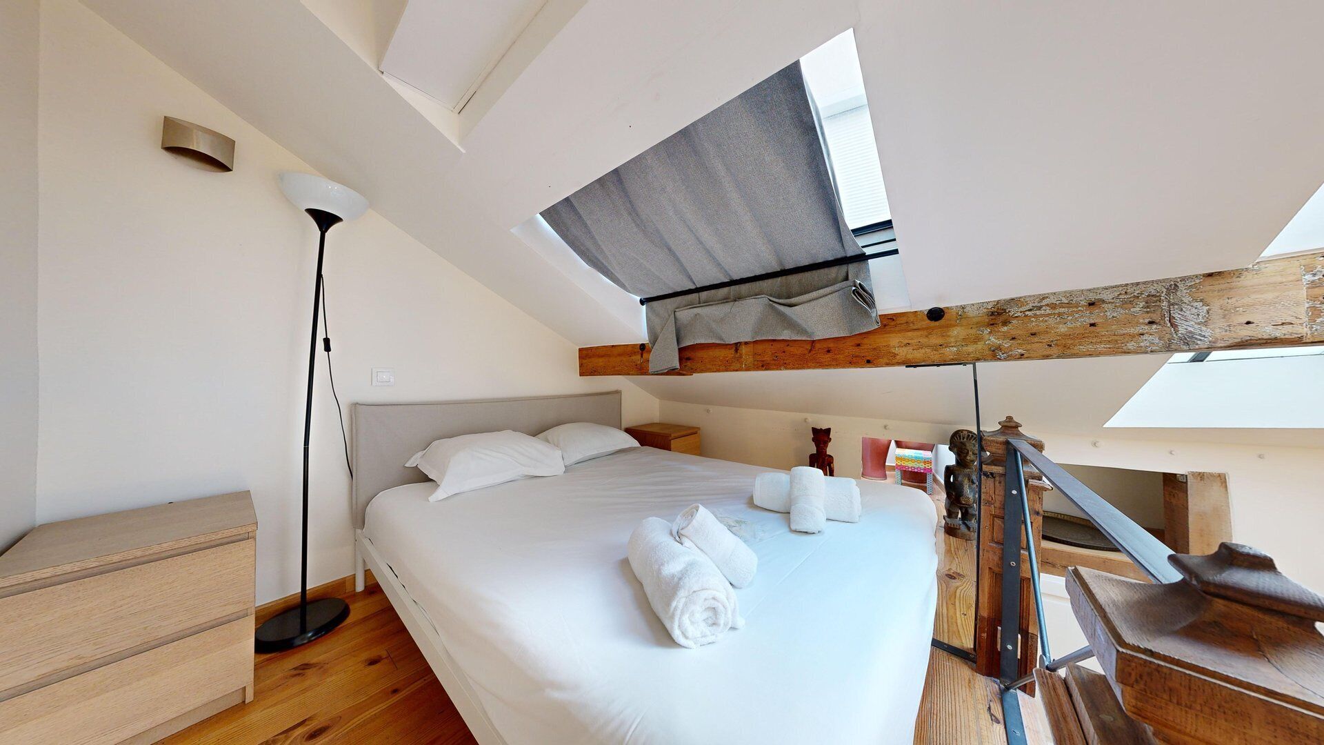Une chambre à coucher avec un grand lit, une table de chevet, une lampe sur pied et une structure en poutres apparentes.