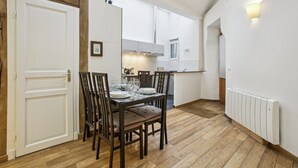 Un comedor con una mesa de madera dispuesta para cuatro personas, rodeada de sillas. Se ve una cocina al fondo con armarios blancos y una ventana.