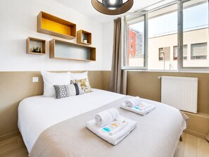 A hotel room with a bed, two side tables, a window with a view of buildings, and a radiator.