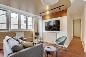 Ein modernes Wohnzimmer mit einer Couch, einem Esstisch mit Frühstücksgebäck, einem an der Wand montierten Fernseher und einem großen Fenster.
