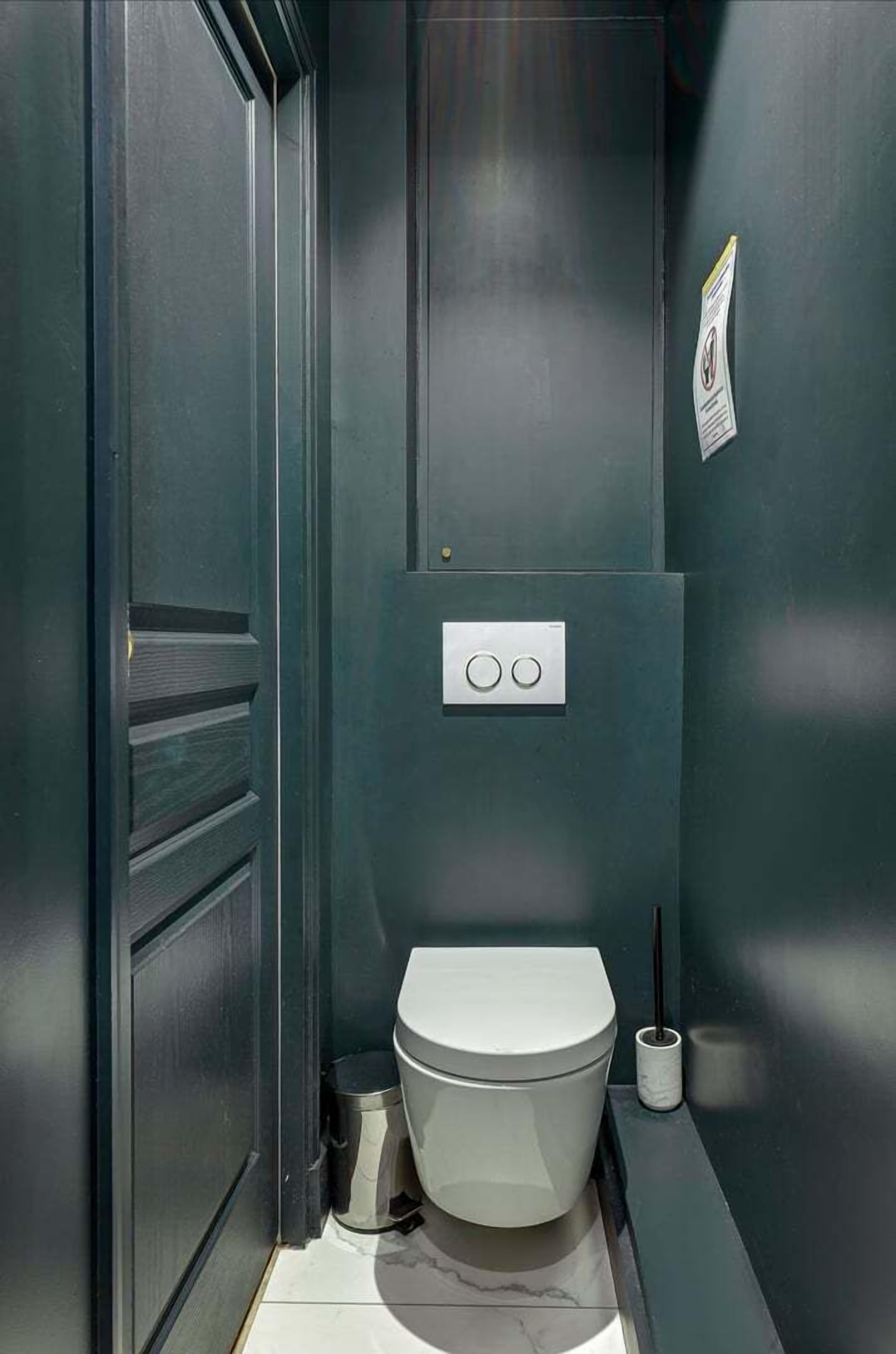 A compact, modern bathroom with a dark color scheme, a toilet, a trash bin, and a wall-mounted flush handle.