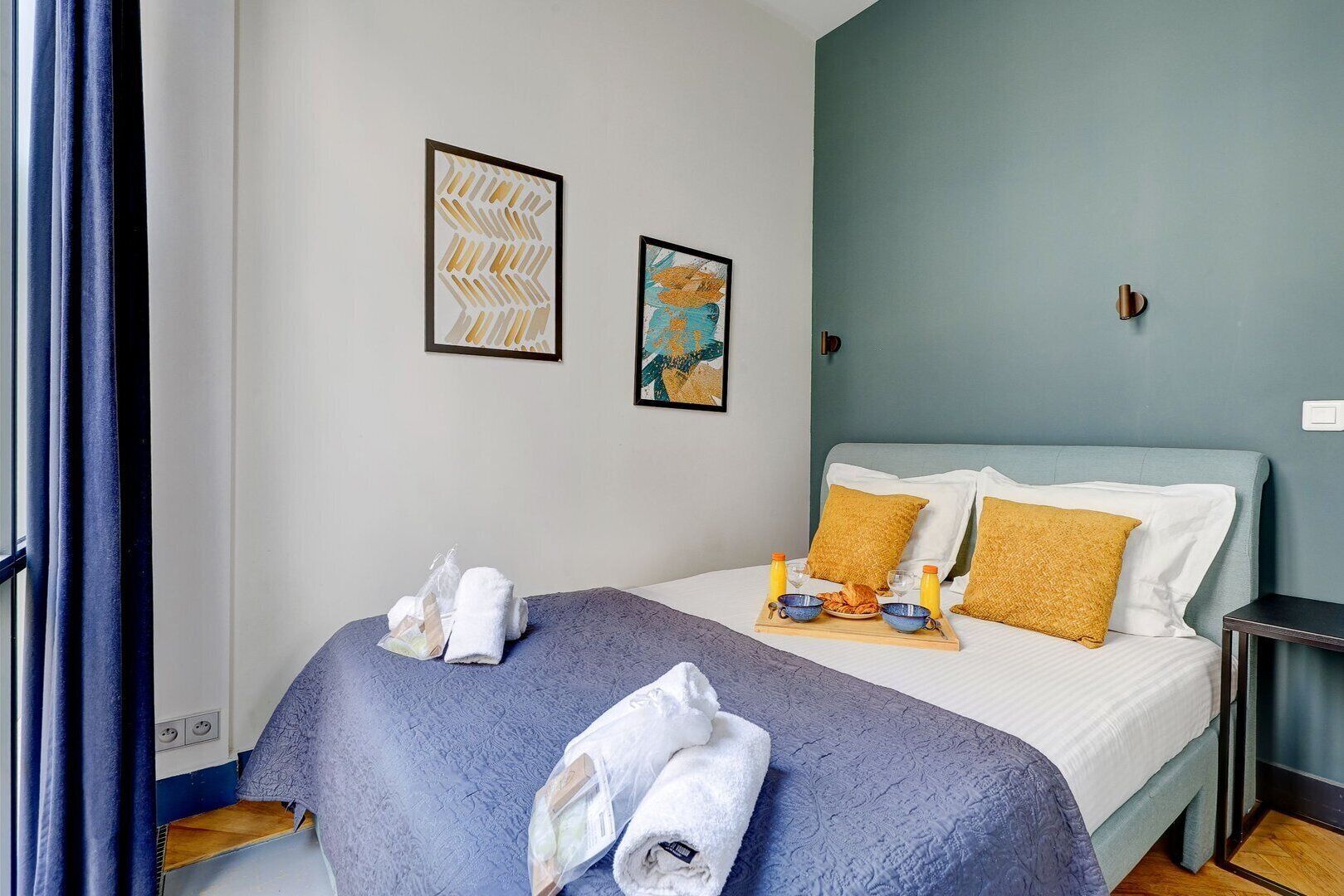 A hotel room with a bed, towels, breakfast tray, and two framed artworks on the wall.