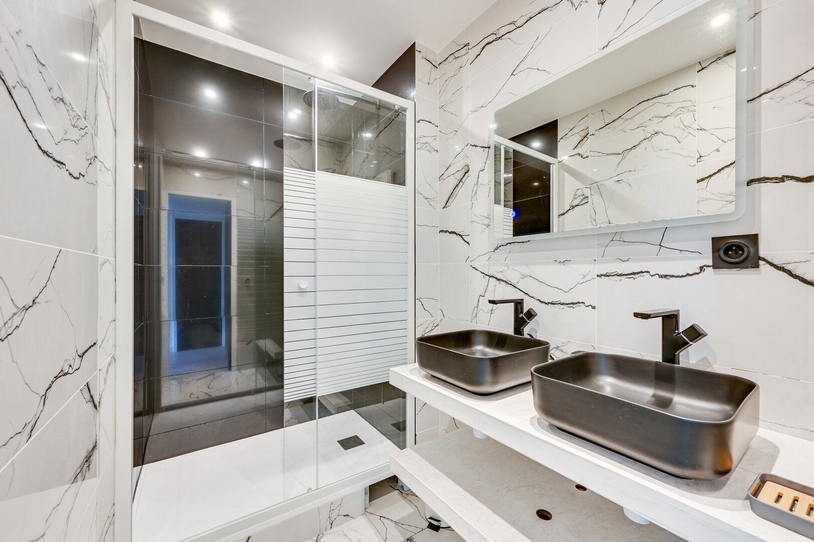 A modern bathroom with marble walls, a glass shower enclosure, and two stainless steel sinks.