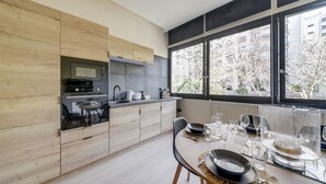 A modern kitchen with a built-in oven and microwave, a dining table set for four with glasses and plates, and large windows offering a view of trees and buildings.