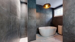 A modern bathroom with a freestanding bathtub, dark stone walls, and a large window providing natural light.